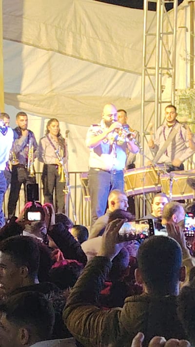 The image captures a vibrant outdoor musical performance by a band on a brightly lit stage, with an engaged audience in the foreground. The scene appears to be taking place in the evening or at night, indicated by the artificial lighting illuminating the performers against a dark background.

At the center of the stage, a bald man with a beard is actively playing a trumpet. He is wearing a light-colored, short-sleeved shirt with epaulets and dark trousers. To his left, another man with a beard holds a trumpet, and further left, a woman with long hair also plays a trumpet. To the right of the central trumpet player, two men are performing on drums, which have purple accents. The stage itself is set up with a white canvas or tent-like material forming the backdrop and ceiling, supported by metallic truss structures visible on the right.

In front of the stage, a crowd of people gathers, watching the performance. Many audience members are holding up smartphones, recording the event, with the screens of their phones visibly displaying the stage. The audience appears to be standing close together, indicating a lively atmosphere. The overall impression is that of a public concert or celebration. The event is taking place in Ash-Shaghour Municipality, Syria. There is no visible text in the image.