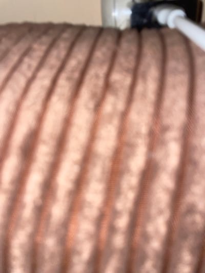 The image shows a close-up, out-of-focus view of a ribbed, brown fabric surface. The ribs run diagonally from the top left to the bottom right. In the upper right corner, a blurred white cable is visible, connected to a dark object, possibly an electrical outlet or adapter. The lighting appears to be from the top and right, casting shadows on the fabric. The image is taken at a low angle, focusing on the texture and color of the material and the partial view of the cable. There are no visible people or identifiable surroundings, making it impossible to determine the specific location within Malevo, Bulgaria.