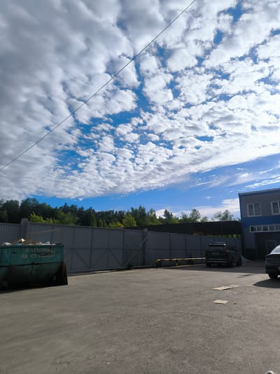 The media file depicts an outdoor scene on a sunny day with scattered clouds. The foreground shows a paved area, possibly a parking lot or industrial yard, with a large green dumpster filled with debris on the left. A grey metal fence stretches across the middle ground, separating the yard from a line of trees behind it.

On the right side of the frame, a modern building with a blue and grey facade is visible. Several cars are parked in the yard, including a dark SUV with a roof rack and another car partially visible on the far right. A yellow and black striped barrier arm is also present.

The sky is a prominent feature, filled with fluffy white clouds against a vibrant blue background, suggesting fair weather. A thin wire or cable runs diagonally across the upper portion of the image. The overall impression is one of an industrial or commercial area on a pleasant day.

Visible text on the green dumpster reads "МАШИНЫ" and "НЕ СТАРИ" which translates to "MACHINES" and "NOT OLD".

The location context is Korolyov, Russia.