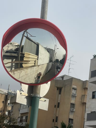 A convex mirror with a red frame reflects a street scene with corrugated metal structures and a sidewalk where a person lies down. In the background, multi-story buildings rise against a bright sky. The mirror is attached to a green pole. Adjacent to the pole is a building. An antenna is visible on top of the building in the background.
