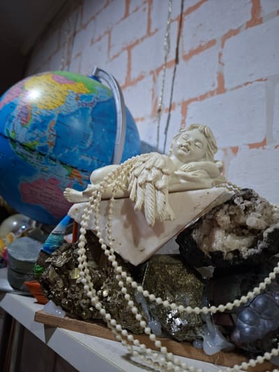 The media file features a close-up, slightly angled shot of several objects arranged together. In the foreground, a small, light-colored figurine of a winged child, possibly an angel, is draped over a triangular white object. The figurine has its eyes closed and a serene expression. Strands of small, white beads, resembling pearls, hang loosely around the figurine and the other objects.
Behind the figurine, a collection of mineral specimens is visible. These include a large piece of glittering, golden pyrite, as well as darker, rougher rocks with crystalline formations. White glue or adhesive is used to hold some of these specimens together and to the shelving.
To the left of the figurine and minerals is a portion of a globe, showing continents and oceans in various colors. The globe appears to be a standard desk model. The background consists of a ceiling with exposed brickwork, painted white and with visible orange lines marking the grout. The lighting suggests an indoor setting, likely artificial.
There are no people or explicit activities taking place. The scene is a static arrangement of decorative and mineral items. The overall impression is one of a display or collection, possibly a personal curation of objects. No text is clearly visible in the image. The location context of Ridder, Kazakhstan, provides a geographical setting for this collection.