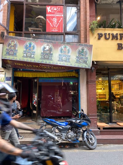 In Kathmandu Metropolitan City, Nepal, a storefront for "SURENDRA'S TIBETAN THANKA TREASURE" is depicted. The shop features a glass entrance with red-tinted glass and a sign above the entrance that reads "TIBETAN THANKA TREASURE." A colorful banner with Buddhist deities in seated poses hangs above the shop entrance. To the right of this storefront, another establishment with a yellow facade has the partially visible word "PUMPKIN" in raised letters. A blue and black motorcycle is parked on the street in front of the Tibetan shop. In the foreground, two individuals on a motorcycle are out of focus due to motion blur, suggesting movement. The street is paved, and there are steps leading up to the entrance of the Tibetan shop. The overall environment appears to be a busy urban street.