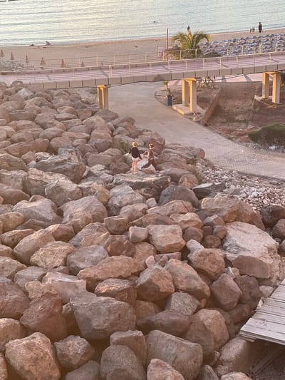 This is an outdoor shot taken in Mogán, Spain. It shows a beach with loungers and umbrellas, a concrete walkway with railings, and a large pile of rocks. Two people are walking on the rocks, while others are on the beach. A palm tree is visible. The lighting suggests either sunrise or sunset. The image is taken from an elevated angle.