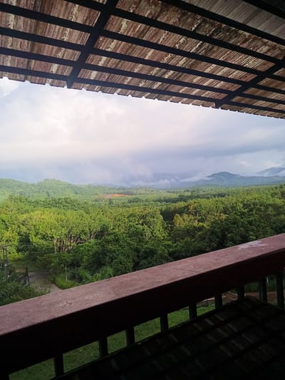 The image shows a view from a balcony in Ban Tak Daet, Thailand. The balcony has a brown wooden railing and a covered roof with a corrugated texture. Beyond the balcony, there is a lush green landscape with trees and hills in the distance. The sky is overcast with cloudy weather conditions. There are no visible people in the frame. The scene conveys a sense of peaceful seclusion in a natural setting.