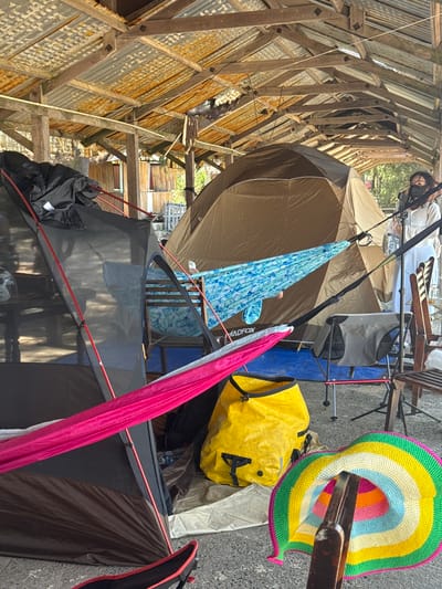 The image depicts an outdoor camping setup under a rustic, open-sided shelter with a corrugated metal roof supported by wooden beams. Several tents are erected within the shelter. In the foreground, a dark-colored tent is partially visible, with bright pink and blue hammocks strung between it and other unseen supports. A large yellow dry bag sits on the ground next to the tents. To the right, a person wearing a mask and white top stands near a larger, tan-colored tent. Wooden chairs are placed around the area, and a colorful crocheted item, possibly a hat or a mat, is lying on the ground in the lower right corner. The environment appears somewhat unkempt, with visible wear and tear on the shelter's structure, suggesting a potentially remote or informal camping location. The backdrop shows vegetation and what appears to be a wooden building, hinting at a rural or semi-rural setting. While the specific location in the city of Thuận Thành, Vietnam is not directly identifiable from visual cues, the scene suggests an outdoor recreational area.