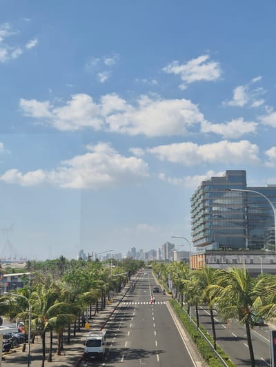 The image depicts a multi-lane road running parallel to the coast, lined with palm trees and buildings in Pasay, Philippines. A tall, modern glass building is on the right side of the frame. A clear, blue sky with fluffy clouds occupies the left half of the image. Vehicles can be seen on the road. Pedestrians are walking across the crosswalk.