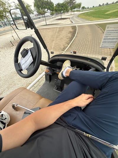 A golf cart interior shows two people riding on a golf course. One person is driving, wearing navy pants and white tennis shoes, with their foot on the gas pedal. The other person is sitting next to them, wearing a black shirt. A golf club rests between the two passengers. The golf course is visible through the windshield, with a paved path and green grass. A scorecard is attached to the steering wheel.