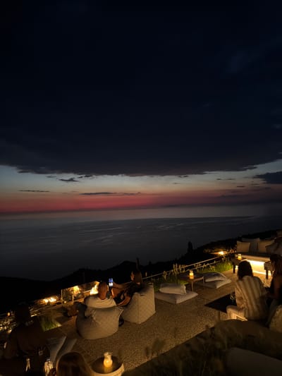 The image depicts an outdoor seating area at dusk with a panoramic view of the sea. The sky displays a gradient of dark blue to red, indicating sunset. Several people are seated on beanbag chairs and a couch, engaged in conversation or looking at their phones. A fire pit is visible, providing warmth and light. The seating area is decorated with cushions and small tables with candles. The location appears to be on a hillside, with vegetation and the sea stretching out to the horizon. The overall atmosphere is relaxed and social.