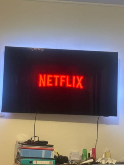 The image shows a television screen displaying the Netflix logo in red. The TV is mounted on a white wall. A shelf with various objects is mounted on the wall to the right of the TV. The location cannot be confirmed to be in Waldmünchen, Germany.