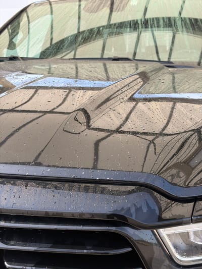 This close-up, angled photograph, taken in Bezborodovo, Russia, captures the side and front-right section of a dark-colored car, likely black or dark grey. The car's body panels and grille are covered in numerous water droplets and streaks, indicating it has recently been washed or exposed to rain.
The shiny surface of the car vividly reflects its surroundings. On the left and central portions of the image, prominent reflections reveal a geometric pattern, suggesting a building's glass facade, an awning, or a structured overhead cover with visible beams or frames, set against a bright, possibly sky-lit background. These reflections are slightly distorted by the water and the car's contours, including a subtle outline of a door handle on the side panel.
To the right, part of the car's front features are visible, including vertical slats of the grille and a bright segment of a headlight. The grille also shows water droplets, some mixed with fine particulate matter or dirt. The overall lighting is bright, indicative of daytime. There is no visible text in the image.