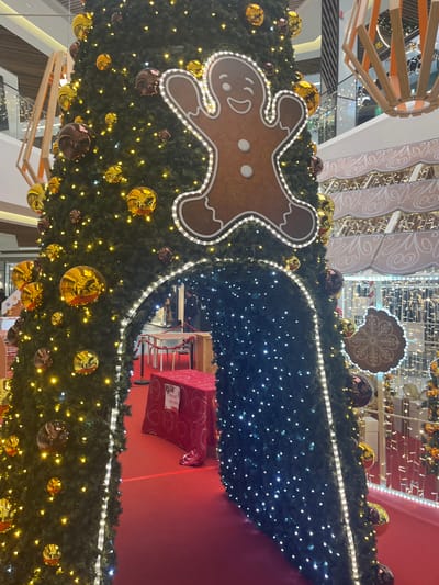 A large Christmas display with a gingerbread man adorns a garland decorated with gold and brown ornaments. A section of the display features a dark green, densely lit area with tiny white lights. A red table with a patterned tablecloth sits below the dark green area. The display is surrounded by architectural elements, including wooden beams and a white, patterned structure. The scene is festive and holiday-themed.