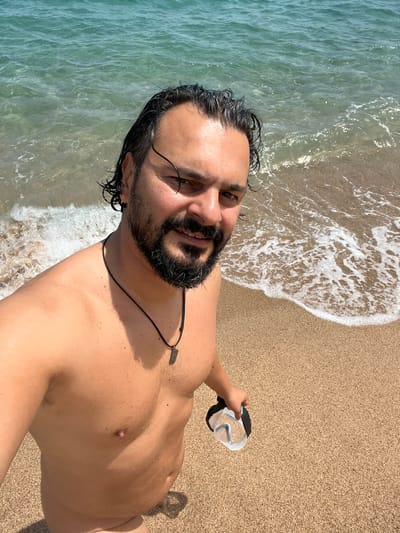 A man with dark hair and a beard stands on a sandy beach, with the ocean in the background. He is wearing a necklace and holding a pair of goggles in his right hand. The water is a clear turquoise color and appears to be shallow. The sand is light brown and fine-grained. The sun is shining, casting a shadow from the man on the sand. The scene appears to be a typical day at the beach in Sant Pol de Mar, Spain.