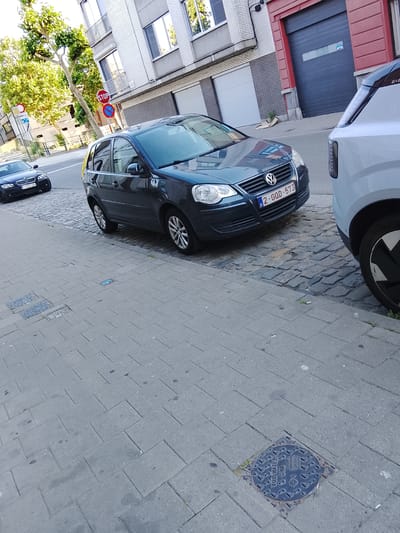 In Ghent, Belgium, on a cobblestone street lined with buildings, a dark blue Volkswagen Polo is parked. The car's license plate reads "2-GOD-573". To its left, a black BMW is also parked. In the background, a large red garage door is visible, and a stop sign is posted on a pole. The street is paved with gray rectangular tiles and cobblestones, and a metal manhole cover with the inscription "VUIL WATER" is present on the sidewalk.