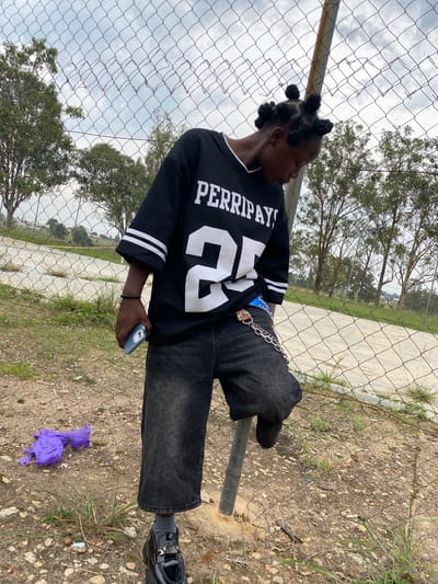 A young person with their hair styled in Bantu knots stands leaning against a chain-link fence in Zawan, Nigeria. The individual is wearing a black oversized jersey with white stripes on the sleeves and "PERRIPAY!" and "25" printed on the front. They have on dark wash, wide-leg jeans and black chunky loafers with grey socks. A silver chain with a blue embellishment is attached to their belt loop. In their right hand, they hold a light blue smartphone, and on their left wrist, they wear a dark beaded bracelet. To the left of the person, on the dusty ground, is a crumpled piece of purple plastic. The background consists of sparse green trees and a cloudy sky. The ground is covered in dirt, small stones, and patches of dry grass.