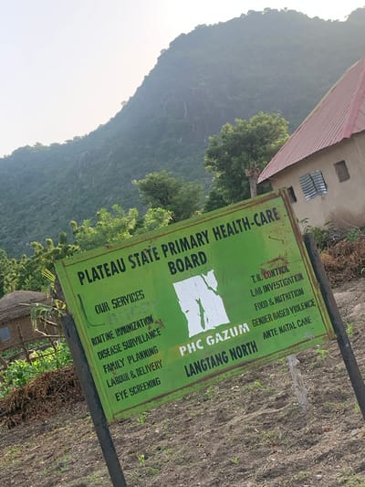 This media file depicts a green sign with white lettering, placed outdoors in what appears to be a rural or semi-rural setting in Reak, Nigeria. The sign is tilted and weathered, suggesting it has been exposed to the elements for some time.

The sign lists services offered by a "PLATEAU STATE PRIMARY HEALTH-CARE BOARD" located at "PHC GAZUM LANGTANG NORTH". Visible services include "T.B. CONTROL", "LAB INVESTIGATION", "FOOD & NUTRITION", "GENDER BASED VIOLENCE", "ROUTINE IMMUNIZATION", "DISEASE SURVEILLANCE", "FAMILY PLANNING", "LABOUR & DELIVERY", "ANTE NATAL CARE", and "EYE SCREENING". The backdrop features a building with a red corrugated roof and some windows, as well as lush green trees and a steep, forested hillside. The sky is bright, indicating it is daytime, and the overall lighting suggests a potentially hazy or overcast condition. The ground appears to be dirt with some sparse vegetation. The scene conveys a sense of a community health service in a developing area. No people are visible in the image.