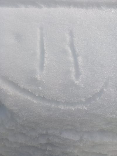 The image displays a close-up, downward-angled view of a snow-covered surface in the city of Яромаска, Russia. The snow is white and granular, exhibiting varied texture and subtle shadows from natural light. Etched into the upper-middle section of the snow is a simplified smiley face, formed by three distinct, shallow indentations: two parallel vertical lines serving as eyes, and a single upward-curving line beneath them acting as a mouth. The carvings are precise, creating clear depressions against the slightly raised snow surface. The lower portion of the image reveals more pronounced undulations and layering within the snow, suggesting depth or accumulation. No individuals, animals, or man-made objects other than the snow etching are present in the scene. The overall environment indicates outdoor winter conditions.