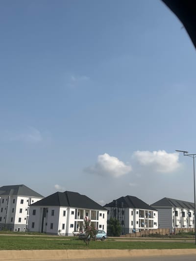 This image depicts a row of modern, multi-story residential buildings with white facades and dark tiled roofs, situated in the city of Karum, Nigeria. The buildings appear to be recently constructed or undergoing finishing touches, as evidenced by the presence of various architectural features like balconies and multiple windows. A light blue vehicle is parked in front of one of the buildings. The foreground shows a well-maintained green lawn bordered by a concrete barrier, suggesting a planned residential development. The sky is clear with a few scattered white clouds. A streetlamp is visible on the right side of the frame. The overall scene suggests a quiet, possibly suburban, residential area.