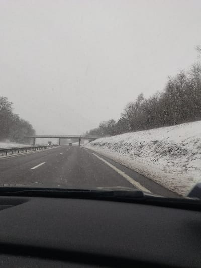 This image captures a snowy highway scene near Crusnes, France, viewed from inside a vehicle looking forward through the windshield. Light snow is falling, creating a hazy, overcast gray sky and a diffuse, low-visibility environment. The multi-lane asphalt highway, appearing wet with light snow accumulation, extends into the distance, marked by a dashed white lane divider and a solid white line on the right shoulder. To the immediate right, a snow-dusted embankment rises, densely covered with bare deciduous trees showing patches of brown earth. Trees also line the distant left side of the road. An overpass, a gray bridge structure, spans the highway in the mid-ground. Two vehicles are visible on the highway ahead: a light-colored car approaching the overpass, and a darker, possibly larger vehicle further back beneath the overpass. The dark gray dashboard and the base of the windshield of the observer's vehicle occupy the bottom foreground. No individuals are discernible outside of vehicles.