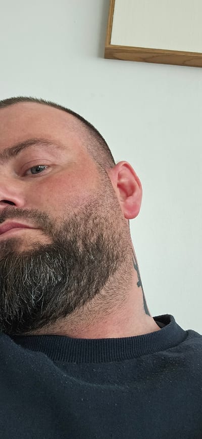 The image shows a close-up of a man's face, likely taken indoors. The man has a beard with graying hairs, fair skin, and a blue eye visible. He is wearing a dark-colored sweater. A tattoo is visible on his neck. The background consists of a plain white wall with a framed artwork or mirror partially visible in the lower right corner. There is no information given that suggests that the location is in Gütersloh, Germany.