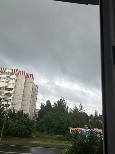 The image depicts a cloudy, overcast sky over a residential area in Sof'ino, Russia. In the foreground, a wet asphalt road with white lane markings is visible. Adjacent to the road is a sidewalk and a patch of green grass. To the left, a multi-story apartment building with a light-colored facade and red decorative elements at the top stands tall. Trees and lush green foliage are scattered throughout the scene. In the middle ground, a commercial building with bright neon signs is partially obscured by trees. A small, bright green car is perched on top of this building. Power lines stretch across the sky, and a utility pole is visible on the left side of the image. The overall atmosphere is somber due to the heavy cloud cover.
