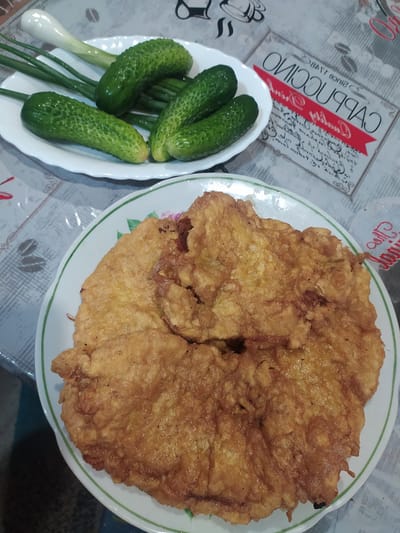 The image shows a plate of food and a bowl of cucumbers on a table. The food on the plate appears to be a large, golden-brown omelet or schnitzel, possibly made from chicken or pork, with a slightly uneven texture. The bowl contains several whole, green cucumbers and some green onions. The table is covered with a patterned tablecloth that has images of coffee cups and the word "CAPPUCCINO" printed on it in a stylized font, with a red ribbon design. The setting appears to be indoors, possibly a home kitchen or dining area in Kobeliachok, Ukraine. There are no people visible in the image. The lighting suggests it could be daytime. The overall impression is of a simple, home-cooked meal.