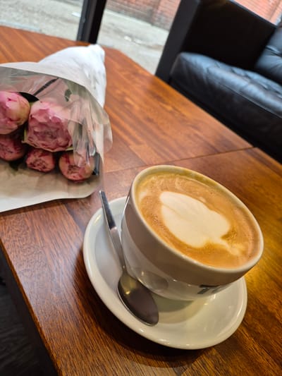 This image captures an intimate indoor setting, likely in West Suffolk, United Kingdom, showcasing a cup of coffee and a small bouquet of flowers on a polished wooden table. The main focus is a white ceramic coffee cup filled with a latte, featuring clear white latte art, resembling a heart, on its frothed surface. A reflective silver spoon rests on the matching white saucer.

Adjacent to the coffee, a delicate bouquet of pink flowers, appearing to be peonies, is bundled in plain white paper and encased in clear plastic wrapping. Several full blooms are visible, contributing a soft, natural element to the scene. These items are positioned on a rich, reddish-brown wooden table, whose distinct grain and polished surface are illuminated by ambient indoor light, possibly enhanced by natural light from a nearby window.

In the upper left background, part of a dark, presumably black, leather sofa with characteristic tufted detailing is visible, suggesting a comfortable seating arrangement. Beyond the sofa, a blurry area hints at an exterior view or an adjacent space, possibly through a window. The overall impression is one of quiet contemplation or a pleasant break. No discernible text is present within the image.