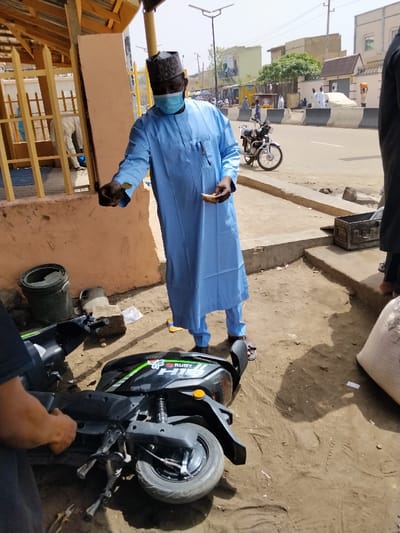 The image depicts an outdoor scene in Fagge D2, Nigeria, under bright daylight. In the foreground, a black and green "RUBY H1S" electric scooter lies on its left side on a dirt ground. A person's arm and hand are visible in the lower-left, positioned near the scooter's front wheel, with pliers visible on the ground.
Standing centrally is an adult male wearing a light blue traditional long-sleeved garment, a dark cap, and a light blue surgical face mask. He holds a dark, crumpled object in his left hand and a lighter-colored, folded item, possibly paper or fabric, in his right hand. He wears dark sandals. Behind him, on the dirt, are a dark green bucket and a lighter, irregular object.
To the immediate right of the central figure, a segment of another person's lower leg, clad in dark trousers and wearing sandals, is visible, standing next to a large white sack on the dirt. A dark, rectangular metal container rests on a raised concrete platform or step.
In the background, a paved road with a central divider separates the foreground area from residential and commercial structures. A silver motorcycle is parked on the far side of the road. Multiple individuals are visible on the road and sidewalks. Buildings of varying heights and colors line the street, with a clear blue sky above. Streetlights with attached solar panels are present along the road. To the left, a light brown structure with yellow wooden lattice elements is partially visible, with another person in white partially visible within it.