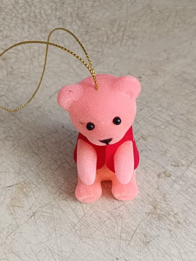 A small, pink, fuzzy teddy bear ornament with a red vest sits on a textured, light-colored surface. The ornament has two black eyes and a small black nose and mouth. A gold string is attached to the top of its head, looping to the left. The background surface appears to be a tabletop or counter with numerous fine, scratched lines and some scattered dark spots. The image was taken at eye-level. There is no indication of the location being in the city of Tebras, Latvia.