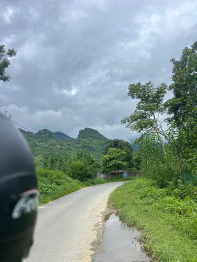 The image captures a scenic view of a winding road in Lùng Tám Commune, Vietnam, under a cloudy sky. In the foreground, the left side of the frame is dominated by the black helmet of a motorcyclist, partially obscuring the view. The road itself is paved and appears wet in places, with a puddle reflecting the overcast sky. Lush green vegetation lines both sides of the road, with dense foliage and taller trees forming a natural barrier. In the background, rolling green hills rise up, shrouded in mist and clouds, their slopes covered in vegetation. The overall atmosphere is one of a rural landscape, possibly captured during or after a rainfall.