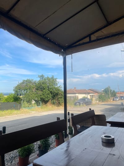 The image presents a static, outward-facing view from a covered seating area in Konstantinovo, Bulgaria, overlooking a semi-rural street and residential landscape under a partly cloudy sky.
In the foreground, a dark wooden table occupies the lower right, featuring a circular metallic ashtray. Partially visible dark wooden benches or chairs are positioned to the left and slightly behind the table. Beneath the benches, on a gravel bed, multiple potted green plants are present. The top portion of the image is framed by a light-colored fabric canopy stretched over a dark metal support structure, indicative of an outdoor shelter. A transparent screen, possibly plastic, separates the interior seating from the exterior view, showing minor imperfections or dust.
Beyond the immediate seating area, an asphalt road traverses the midground. To the left of the road, a strip of dry grass is visible, followed by a dark mesh fence and a large, verdant tree. A decorative green metal gate stands near the tree. Distant, hazy hills are visible on the horizon to the far left.
To the right of the road, several single-story houses with tiled roofs are visible, exhibiting signs of age or disrepair, with some overgrown vegetation around them. A dark-colored SUV is parked in front of one of these houses, partially obscured by foliage. Further down the road on the right, two lighter-colored vans or trucks are partially discernible. Two wooden utility poles with overhead wires stand along the right side of the road.
The sky is bright blue with scattered white cumulus clouds, becoming denser on the right side of the frame. No people or specific actions are depicted within the scene.