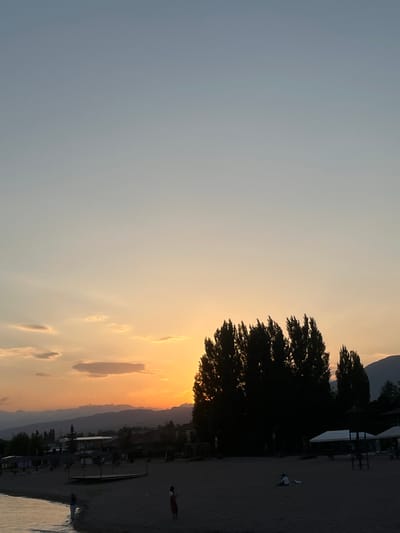 This is a vertically oriented photograph of a sunset over a beach in Choktal, Kyrgyzstan. The sky is a gradient of soft blues and oranges, with wispy clouds catching the last rays of the sun. A silhouetted tree stands prominently on the left side of the frame, its branches reaching towards the colorful sky.
The beach itself is dark and sandy, with several structures that appear to be part of a resort or recreational area. These structures are also silhouetted against the brighter sky. In the middle ground, there are two figures visible on the beach. One appears to be a person in white clothing, possibly lying down or stretching, while another figure in darker clothing is further down the beach, walking away from the camera.
The water of a lake or sea is visible in the lower right corner of the image, reflecting the warm colors of the sunset. The overall mood of the photograph is serene and peaceful, capturing the beauty of a sunset at the end of a day. The time of day is clearly sunset, given the warm colors and soft lighting. There is no visible text in the image.