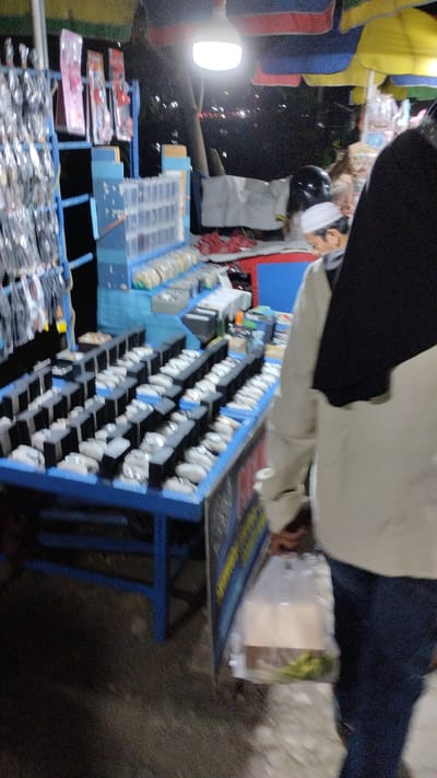 This media file depicts an outdoor stall, likely in Sukodono, Indonesia, at night. The scene is illuminated by a bright, hanging LED light, suggesting a street market or roadside vendor setup. Several stalls are visible, covered by colorful awnings in yellow, blue, and red.
The primary focus is on a stall displaying a large quantity of watches. They are neatly arranged on blue display stands, with individual watches presented on black and white holders, creating a visually organized display. To the left of the watches, another rack holds various items packaged in clear plastic, possibly accessories. Behind the watches, a blue shelving unit contains rows of small, clear plastic boxes, some filled with what appear to be small trinkets or other goods.
Two individuals are partially visible in the frame. One, in the foreground, is wearing a black hijab and a light-colored jacket over blue jeans. They are walking away from the camera, carrying a transparent plastic bag containing some items, including what looks like green produce at the bottom. Further back, another person is wearing a white peci (a traditional cap) and a light-colored garment. They appear to be looking down towards the stall.
The overall impression is that of a bustling night market or a vendor operating late into the evening. The lighting creates a somewhat dim but functional atmosphere, highlighting the merchandise. There is no visible text in the image. The setting suggests a commercial area where goods are sold to passersby.