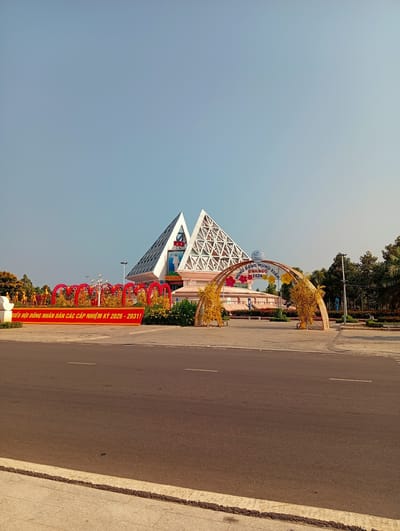 This outdoor image depicts a public space in Phan Rang – Tháp Chàm, Vietnam, under a clear, bright blue sky, suggesting daytime with good weather. The central focus is a modern building, identified by a sign as a Provincial Museum ("BẢO TÀNG TỈNH KH"), featuring two striking white, pyramid-like structures with intricate geometric latticework on their facades.
In front of the building, festive decorations adorn the area. On the left, a row of red, heart-shaped arches is embellished with golden figures and yellow foliage, creating a celebratory pathway. Below these arches, a prominent red banner displays Vietnamese text: "BIỂU HỘI ĐỒNG NHÂN DÂN CÁC CẤP NHIỆM KỲ 2026 - 2031!" (Congress of People's Councils at all levels for the term 2026 - 2031!).
On the right, a large golden arch, possibly crafted from bamboo, is decorated with colorful flower cutouts in pink, yellow, and blue. Above this arch, a circular sign reads: "KHÁNH HÒA MỪNG ĐẢNG MỪNG XUÂN BÌNH NGỌ 2026" (Khánh Hòa – Celebrating the Party, Celebrating Spring, Year of the Horse 2026). These decorations and visible dates suggest preparations for or ongoing celebrations of the Lunar New Year (Tết) in 2026, alongside a civic or political event.
The foreground shows an empty asphalt road and a paved sidewalk. Trees with both green and yellow foliage line the sides of the area, framing the festive scene.
