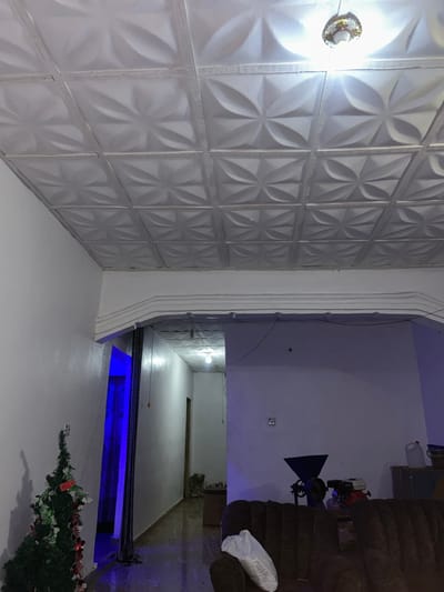 A room with textured white wall panels and a ceiling light is adjacent to another room through an arched doorway. The second room contains a brown sofa, lighting equipment, and miscellaneous items. Further down, the room extends into a blue-lit area with a Christmas tree in the corner. The spaces appear to be brightly lit, creating a sterile and modern aesthetic.