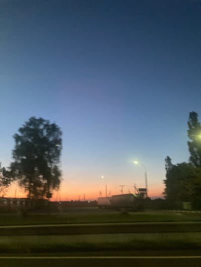 This image depicts a twilight scene likely captured from a moving vehicle on a road in the vicinity of Ракітніцкі сельскі Савет, Belarus. The sky exhibits a gradient of colors, transitioning from a deep blue at the top to hues of orange and pink near the horizon, indicative of sunset or sunrise. In the foreground, a dark, blurred strip suggests a road surface. To the left, a large, heavily blurred tree stands prominently. Further in the middle ground, various indistinct shapes of buildings, poles, and what appear to be trucks or trailers are visible, illuminated by streetlights. To the right, several tall, blurred trees and streetlights punctuate the landscape. The overall impression is one of a rural or semi-urban environment at dusk, characterized by motion blur due to the likely movement of the camera.