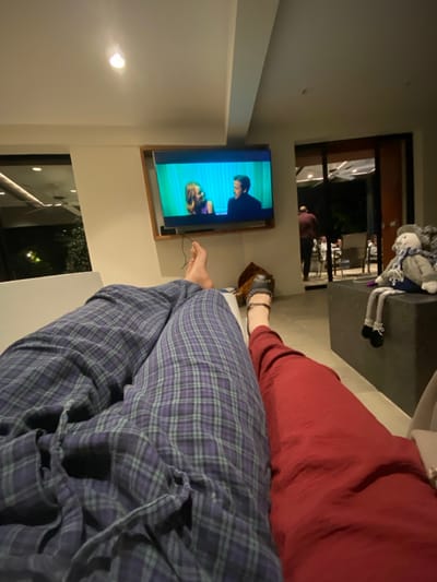 A person wearing plaid pajama pants is lounging in front of a television, which displays a scene from a show or movie. Another person wearing a red garment sits beside them. A doll sits on a surface near the television. Through a window, greenery and exterior lights are visible, while the interior features a modern design.