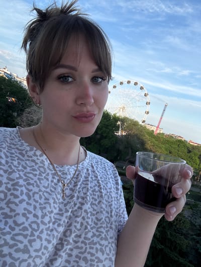 A person is holding a clear plastic cup with a dark liquid. The subject has fair skin, shoulder-length brown hair with bangs, and is wearing a white and grey leopard-print shirt with a gold necklace with a cross pendant. In the background, a large Ferris wheel and a tall drop tower amusement ride are visible. Behind these structures is a line of green trees and buildings. The sky is blue with scattered clouds. The location is Yevpatoriya, Ukraine, based on the distinct architecture and presence of these specific amusement park attractions.