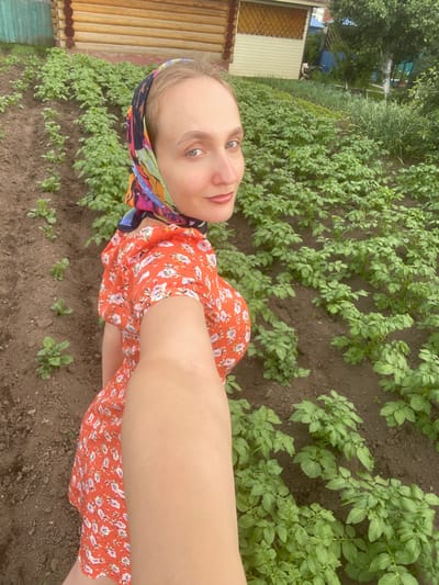 In Kuyeda, Russia, a young woman with blonde hair is captured in a selfie-style photograph taken outdoors in a garden. She is wearing a vibrant orange dress with a floral pattern and a colorful headscarf. The background reveals rows of lush green potato plants stretching into the distance, interspersed with dark soil. A wooden house with a light-colored trim is visible on the left side of the frame, partially obscured by a green fence. Trees and other vegetation can be seen further in the background, suggesting a rural or suburban setting. The woman is looking directly at the camera with a slight smile.