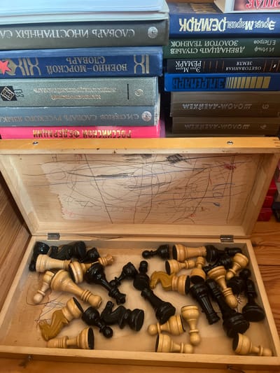 The image shows an open wooden box containing a collection of black and white chess pieces scattered inside. Some of the chess pieces are lying on their sides, while others are standing upright. The inside of the box lid is covered in child-like scribbles made with pencils and crayons. To the left of the open chess box, a stack of books is visible. The spines of these books are mostly in Russian, with titles such as "СЛОВАРЬ ИНОСТРАННЫХ СЛ" (Foreign Words Dictionary), "ВОЕННО-МОРСКОЙ СЛОВАРЬ" (Naval Dictionary), and titles by authors like Erich Maria Remarque and Sholem Aleichem. The setting appears to be indoors, possibly a shelf or desk, with wooden paneling in the background. The overall impression is one of domestic clutter and childhood play.