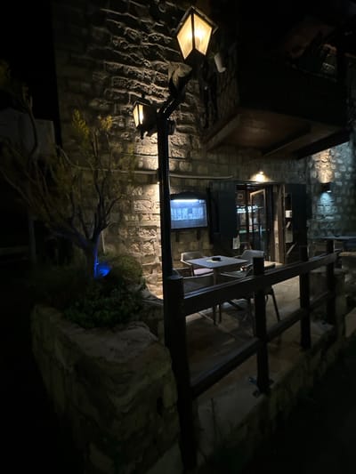 The image depicts an exterior view of a restaurant or cafe at night, likely located in Peyia, Cyprus. The building is constructed of stone. To the left, a small tree is illuminated with blue light, surrounded by greenery. A lamppost with a hexagonal lantern stands near the tree.
In the foreground, a wooden railing separates the viewer from a seating area. This area contains several tables and chairs, some of which are visible through the railing. One table has an ashtray on it.
Further back, a doorway leads into the establishment. The door is open, revealing a dimly lit interior with more tables and chairs. Above the doorway, a sign with lettering is visible, though it is too blurry to read. To the left of the doorway, a backlit display screen shows what appears to be a menu or some form of visual content. Shutters are positioned on either side of the entrance. The overall ambiance is dark and atmospheric, illuminated by the artificial lights of the building and the street lamps.