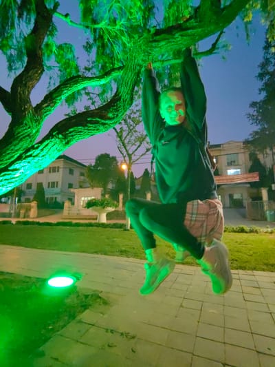 The image depicts an outdoor night scene in an urban park or public space within Sa Pa, Vietnam. A person is observed suspended from a thick, gnarled tree branch, gripping it with both hands. The individual wears a dark long-sleeved shirt, dark leggings, light-colored plaid shorts layered over the leggings, and light-colored athletic shoes. Their face, illuminated by green light, shows a discernible smile.
The tree and its foliage are intensely illuminated by green light, emanating from visible ground-level uplights embedded in the adjacent paved walkway. The ground consists of square-patterned pavers and a section of grass lawn.
In the background, under a dark blue sky, multiple buildings are visible. To the left, a multi-story white building with several windows is present. Further to the right, another building features a large red banner with Vietnamese text stretched across its facade. An orange-glowing street light is situated near this background building. Manicured bushes and a fence are also visible in the mid-ground.