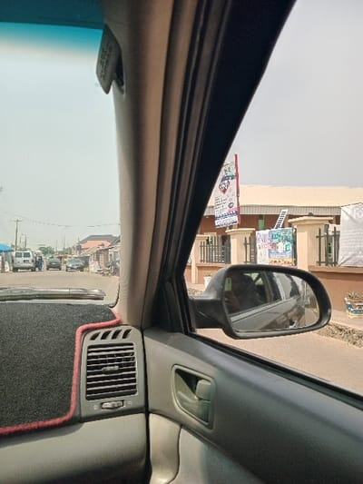 The scene depicts a street in Akobo, Nigeria, observed from the interior of a vehicle. The foreground features a grey dashboard with a black, red-trimmed mat and an air vent. The vehicle's grey A-pillar and the passenger-side door panel, which includes an integrated handle, are prominent on the right. The exterior side mirror reflects a dark vehicle body and a human silhouette.
The exterior view shows a paved road extending into the distance, populated by multiple vehicles including a white van and darker sedans. Structures line the roadway; on the right, a large vertical banner with red, white, and blue elements displays "PRUDENT" and other graphics. A blue sign is affixed to a low wall below it, and another banner partially reads "For" further to the right. Utility poles and power lines are visible on the left. The sky appears hazy or overcast. Indistinct human figures are visible near the white van in the far distance.
