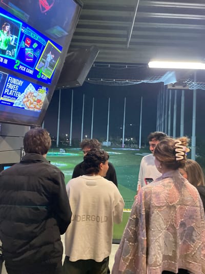 Four people stand at a Topgolf bay, looking at a screen displaying game options and an advertisement for a "Funday Faves Platter" priced at $39. The screen prompts users to sign in, pick a game, and play. One individual is wearing a pink and white patterned shawl, while another wears a white shirt with the word "OIDERGOLD" on the back. A third person is dressed in a black jacket. The Topgolf venue is visible in the background.