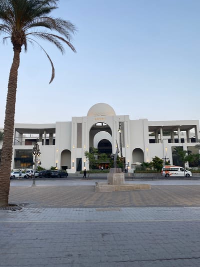 This image captures a modern street scene in Hurghada, Egypt, likely during the late afternoon or early evening under a clear, light blue sky.

The central feature is a contemporary, multi-story building constructed from light-colored (presumably white) materials. Its architecture includes prominent arched openings, balconies, and a central domed section. The word "SERAI" is visible in gold letters on the façade below the dome. In front of the building, a dark, abstract statue or sculpture rests on a stone pedestal.

To the left, a large palm tree with a robust trunk and wide fronds dominates the foreground, extending upwards into the sky. The ground consists of a paved street and sidewalk, laid with interlocking pavers in various patterns. Several vehicles are parked along the street, including multiple dark-colored cars and a white van with orange accents. The van clearly displays the English text "FOX" and "FOX TRAVEL" along with some Arabic script.

A few people are casually walking or standing on the sidewalk, adding a sense of everyday activity to the calm scene. The overall setting suggests a modern resort or commercial area.