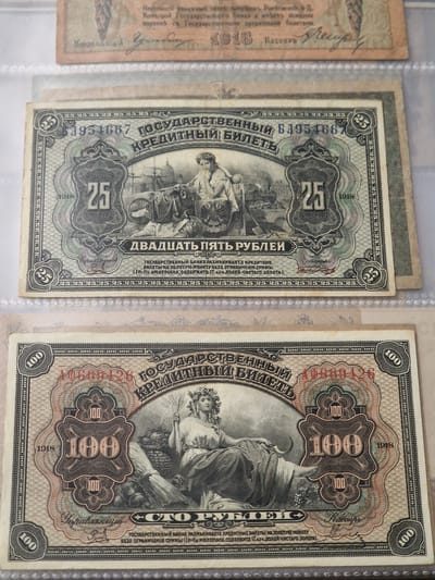 The image displays two Russian Imperial banknotes. The top banknote, dated 1918, is for 25 rubles, and features a classical allegorical scene with a woman holding a sheaf of wheat and a ship in the background. The bottom banknote, also dated 1918, is for 100 rubles, and depicts a similar allegorical figure holding wheat and a cornucopia, with a train and a ship in the background. Both banknotes exhibit intricate designs with ornate borders and serial numbers. The banknotes are presented in a protective clear plastic sleeve. There is no indication or visual element within the image that suggests the city of Voluyak, Bulgaria.
