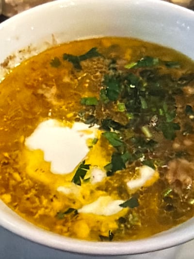 A close-up, top-down, slightly angled shot of a white bowl filled with a steaming, golden-yellow soup. The soup appears to be hearty, with visible pieces of ingredients and flecks of green herbs, likely cilantro, scattered throughout. A dollop of white, creamy substance, possibly yogurt or sour cream, sits prominently in the center of the bowl, swirling slightly into the broth. The broth has an oily sheen, indicating richness. The lighting is soft, suggesting an indoor setting, possibly a kitchen or dining area. The image is somewhat blurry, which can give it an artistic or home-style feel. No text is visible in the image. The overall impression is of a warm, comforting, and flavorful dish.