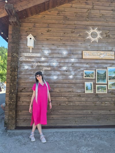 A woman with long, white braided hair and sunglasses stands in front of a wooden wall. She is wearing a bright pink dress and light-colored sneakers with striped socks. The wooden wall has several decorations, including a wooden sun cutout, a birdhouse, and a sign that reads "СЕГОДНЯ Самый лучший день" (Today is the best day). To the right, there is another wooden sign with "КРАСНЫЙ КЛЮЧ" (Krasny Klyuch) and a heart. Below this sign, a collection of framed photographs is mounted on the wall. The scene appears to be outdoors, with a natural landscape visible in the background and a gravel ground in the foreground. The woman is posing for the camera.