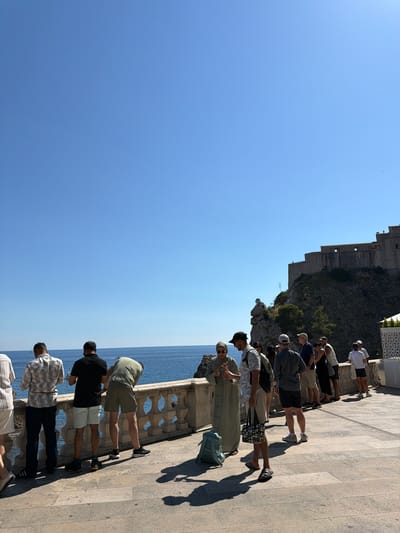 The image depicts a group of tourists standing along a stone promenade overlooking the Adriatic Sea. In the background, a large stone fortress, likely part of the Dubrovnik city walls, is perched on a rocky cliffside. The sky is clear and blue, indicating a sunny day. Several individuals are observing the sea and the fortress, while others are engaged in conversation. One woman is wearing a striped dress and a headscarf, while most of the other people are dressed in casual summer attire. The promenade is paved with stone tiles, and a decorative stone balustrade runs along the edge. The scene conveys a sense of leisure and sightseeing in a historic coastal city.
