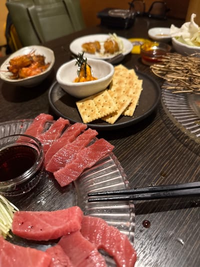 The image depicts a table setting with a variety of Japanese-style food. There are several small bowls containing different appetizers, including what appears to be fried shrimp. A black plate holds crackers and a white bowl contains uni with garnish. Slices of raw tuna are arranged on a glass dish with a small container of dark sauce nearby, next to a set of chopsticks. Based on the presence of Japanese-style food, it's plausible this image was taken at a restaurant in Gimpo-si, South Korea.