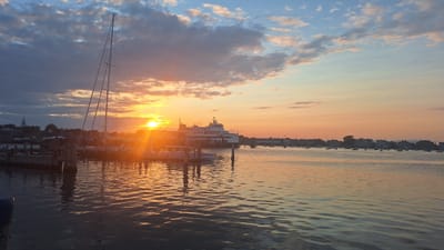 The image depicts a harbor scene in Nantucket, Massachusetts, USA, during sunset. The sun is low on the horizon, casting a warm, golden light across the water and sky. In the foreground, a sailboat with its mast prominently featured is docked on the left side of the frame, with other smaller boats visible in the water. A large ferry is seen in the middle ground, along with a line of buildings forming a townscape on the right side of the harbor. The sky is a blend of blues and oranges, with scattered clouds. The water is calm, reflecting the colors of the sunset and the surrounding structures.